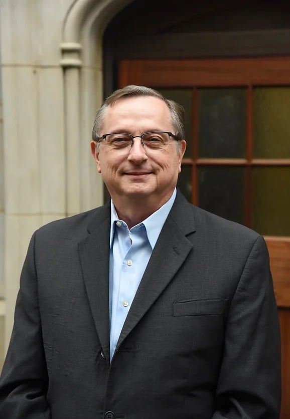 A headshot of a middle-aged man wearing glasses, a black suit jacket, and a pale blue dress shirt.