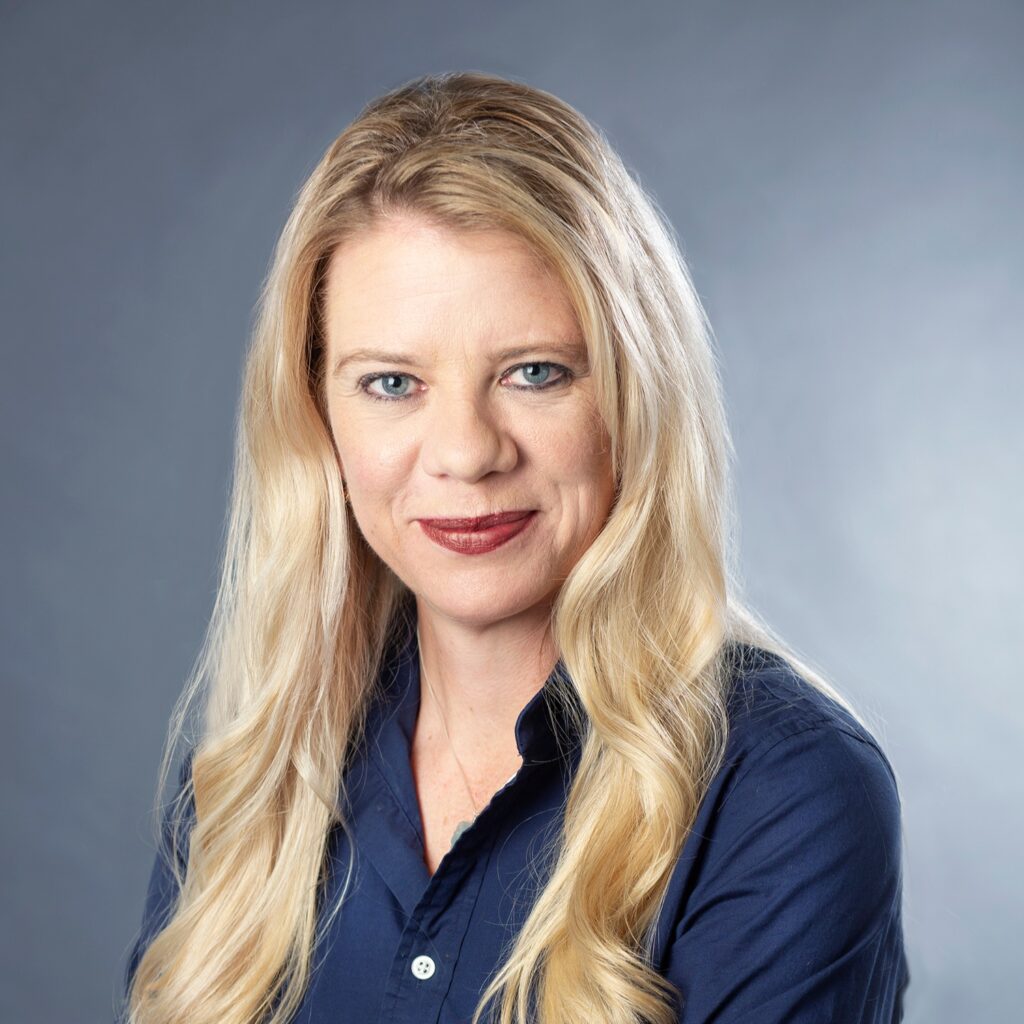 A headshot of a blonde woman, Sandra Leiterman. She is wearing a navy button down shirt and red lipstick.