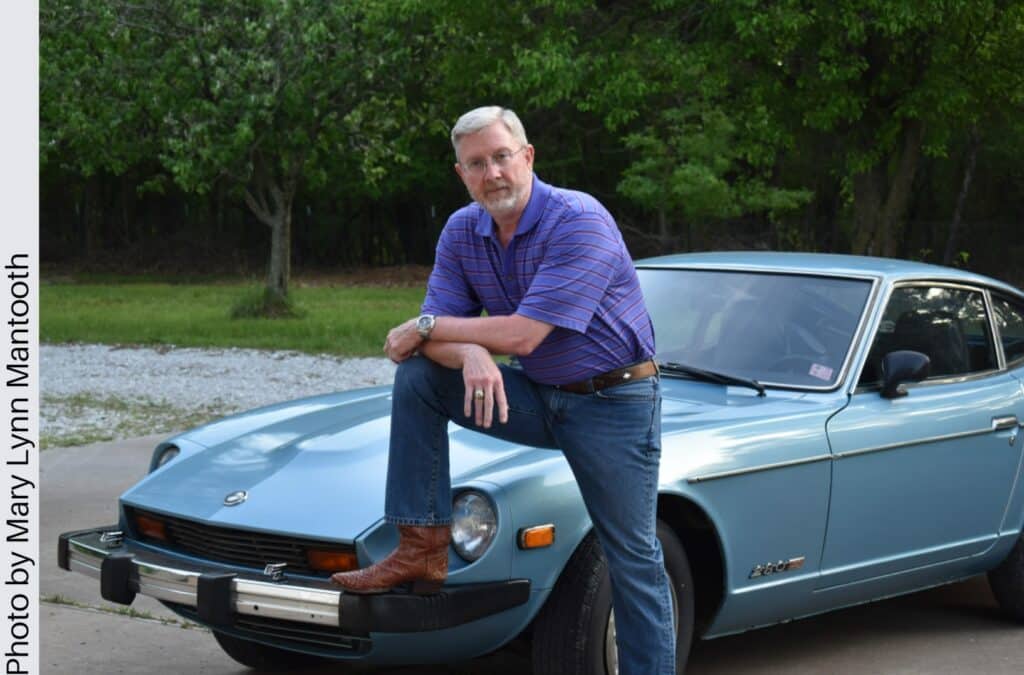 A picture of Dr. Alan Mantooth—an older man in a purple striped shirt, jeans, and cowboy boots with a leg up on an old blue Nissan.