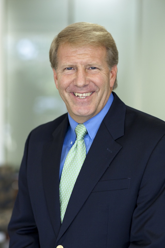 Lonnie Emard headshot: A man with blonde hair, a black suit, blue shirt, and green tie is smiling.