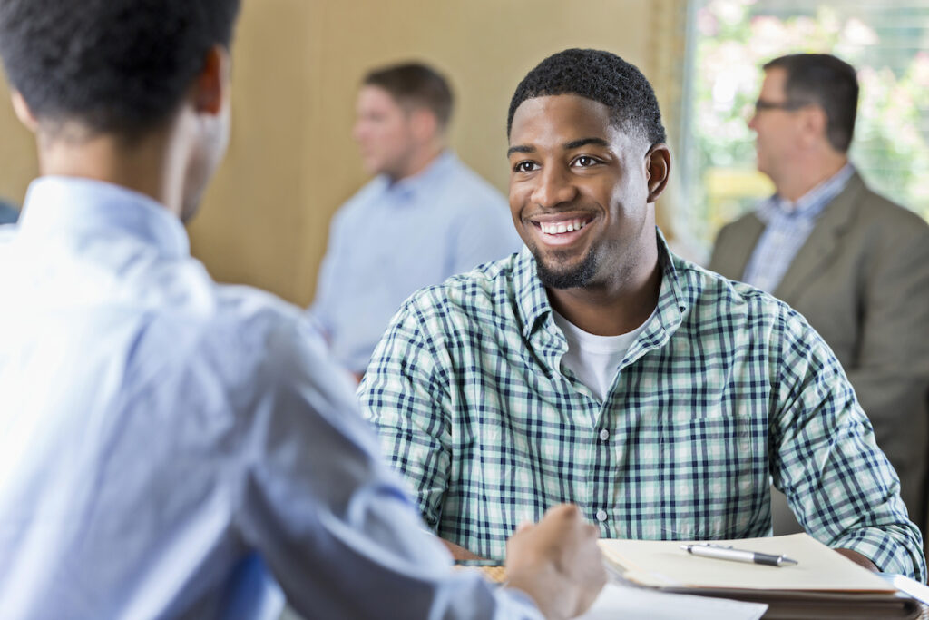 Smiling African American young adult at a job interview in a business center. He is sitting at a desk across from a manager being interviewed for a job. Other applicants are waiting in the background.