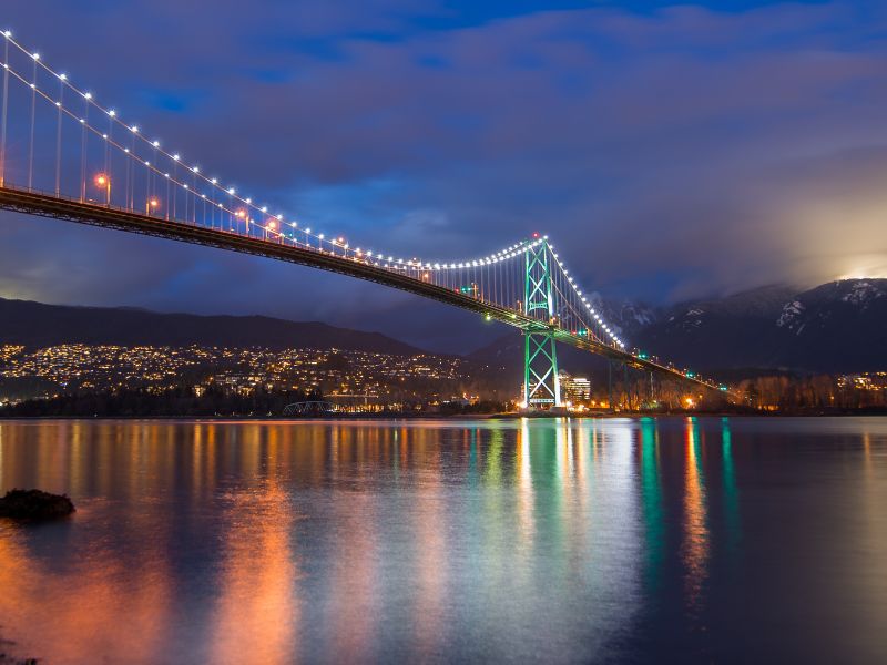 A bridge spanning a wide river at nighttime. City lights and mountains can be seen in the background.