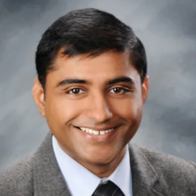 A headshot of an Indian man wearing a gray suit.