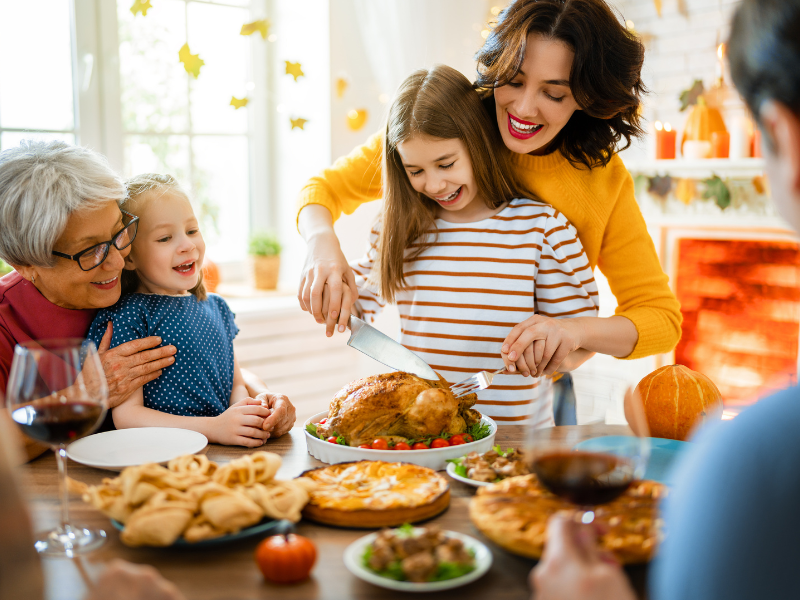 A family celebrating Thanksgiving. A mom holds her daughters hands as they carve the turkey.