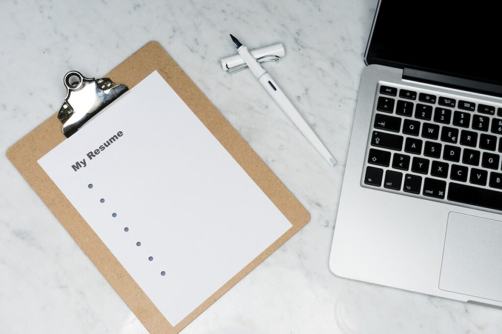 A clipboard with a blank sheet on it that says "My Resume." To the right of the clipboard is a white fountain pen and a silver laptop.