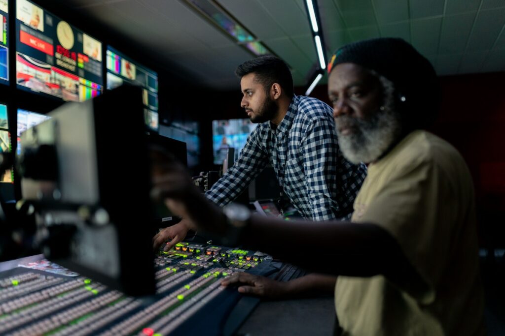 Two men in a dark tech booth looking at the screens and pressing buttons.