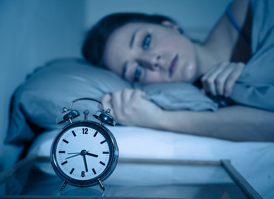 A woman lying on her side in bed staring at her small silver alarm clock.