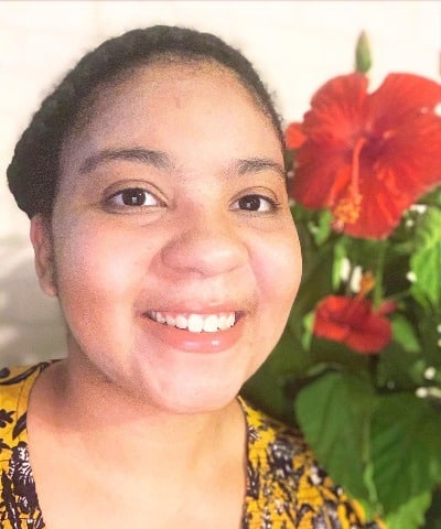 A headshot of Christa Hindman, a black woman with her hair up in a braid crown, wearing a yellow shirt with a Hibiscus plant in the background.