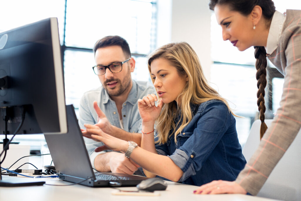A man and woman, both wearing denim shirts, sitting at a desk, gesturing at a laptop. A woman with dark hair in a braid leans over them to talk about giving.