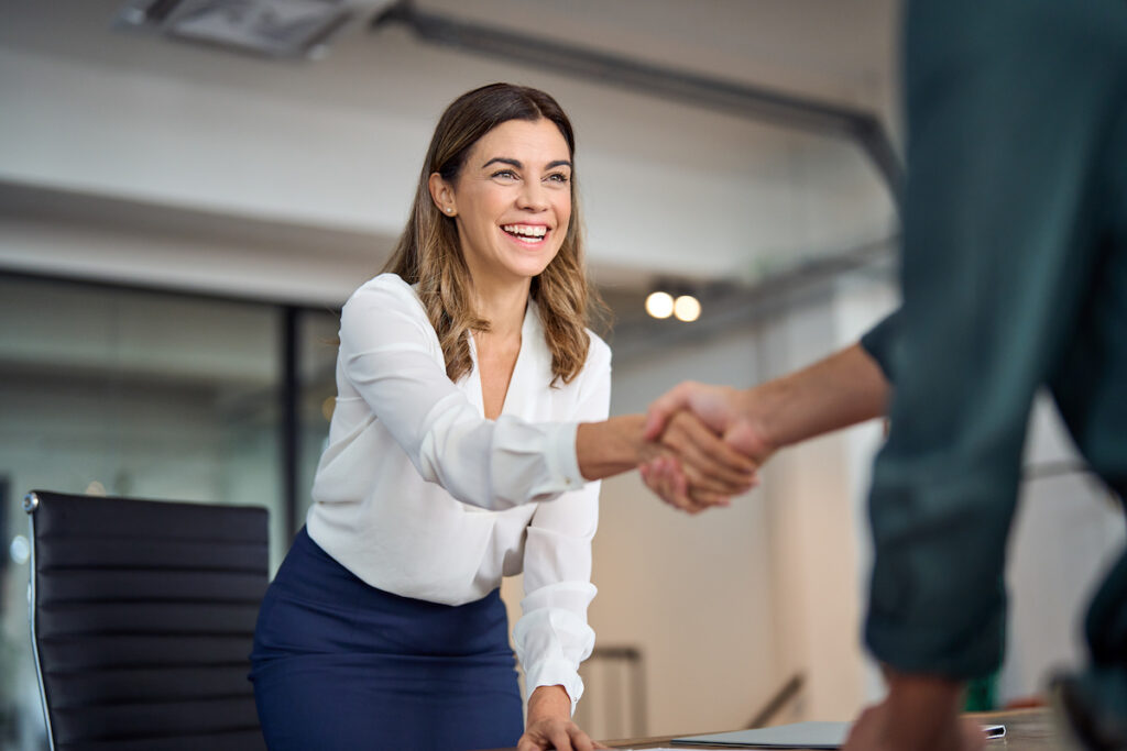 A smiling woman stands from her chair and shakes hands with someone over her desk in the office.