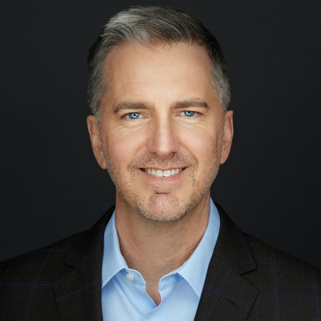 A professional headshot of a man with graying hair and blue eyes wearing a black jacket and blue dress shirt up against a black background.