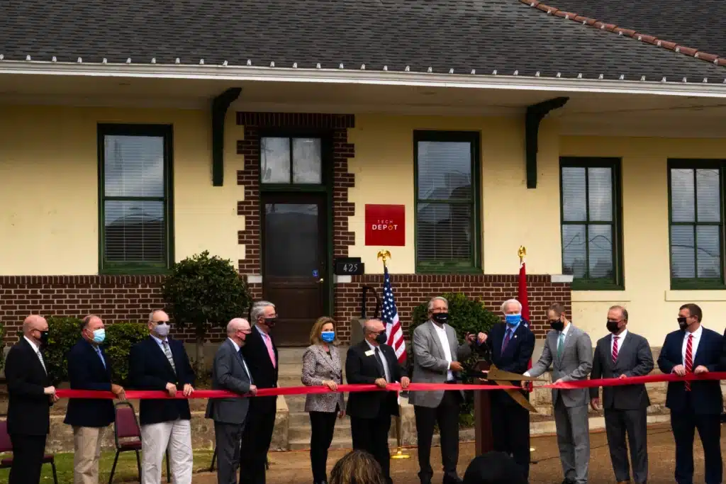 A line of men and one woman in business attire standing and holding a red ribbon in front of the Tech Depot building. Two men in the front hold giant scissors to cut the ribbon. Everyone is wearing a face mask in the picture.