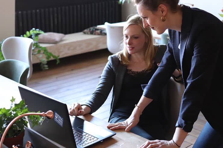 A blonde woman sitting at her laptop in the Apprenticely office while another woman stands over her with both hands placed on the desk, looking at her laptop as well.