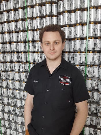 A man standing in front of a wall of beer cans wearing a black "Lost Forty Brewing" polo.