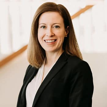 A professional headshot of a middle-aged woman with brown hair wearing a black suit jacket and white blouse smiling at the camera.