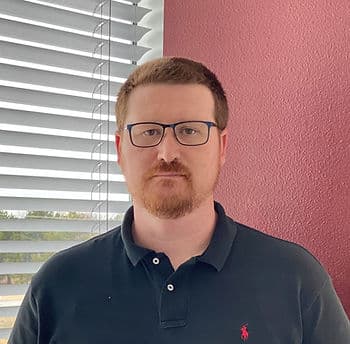 A headshot of a man with red hair and a beard wearing glasses and a black polo standing in a corner with a red wall and a window with blinds behind him.