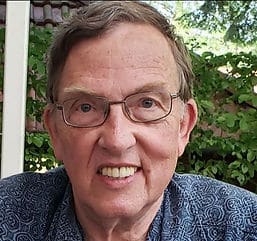 A headshot of an old man with gray hair and glasses wearing a patterned blue shirt with trees in the background.