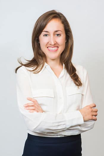 A headshot of a white woman with long brown hair wearing a white button up and black pants, crossing her arms over her stomach.