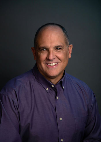 A dark headshot of a balding man wearing a dark purple dress shirt. The background is black and casts shadows.