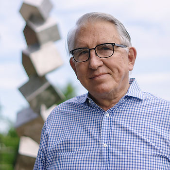 An old man with gray hair and glasses wearing a blue and white checkered shirt standing outside in front of a tall statue made of silver boxes.