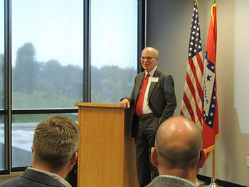 A man in a gray suit and red tie stands beside a brown wooden podium, in front of the US and Arkansas flag.