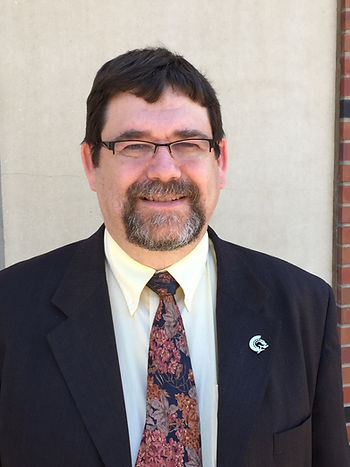 A headshot of a man with black hair and a goatee wearing a black suit jacket with a yellow shirt and floral tie.