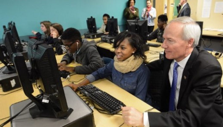 An old man in a suit sitting beside young students in a computer lab class at school.