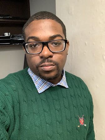 A headshot of a black man with short hair wearing glasses and a green cable-knit sweater.