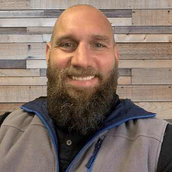 A headshot of a bald man with a full brown beard in front of a wall of multicolored wooden slats. He is wearing a fleece vest and black polo.