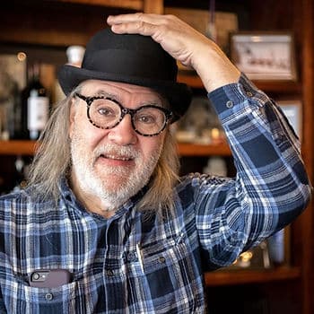 A man with long gray/brown hair and a beard wearing a blue and black plaid shirt. He is wearing a bowler hat and glasses. His iPhone is in his front shirt pocket.