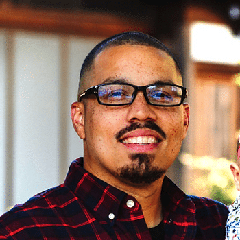 A headshot of a black man with a shaved head and mustache wearing glasses and a black and red plaid shirt.