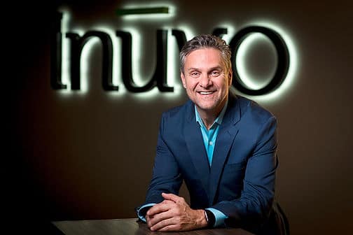 A headshot of a man with gray hair wearing a navy suit and a bright blue shirt standing in front of an Inuvo backlit sign.
