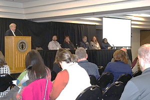 A man standing behind a brown podium next to a panel of 5 people at a black-cloth covered table talking to an audience.