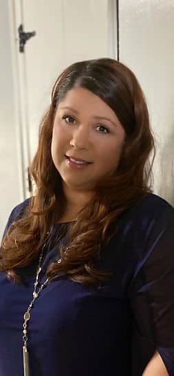 A headshot of a woman with long brown hair wearing a navy shirt and a long silver necklace.