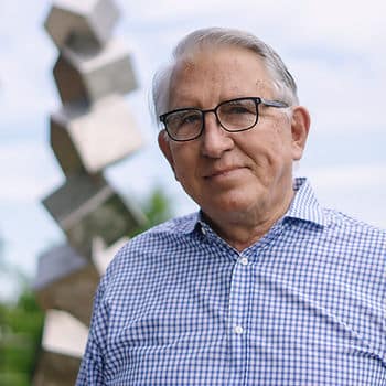 An old man with gray hair and glasses wearing a blue and white checkered shirt standing outside in front of a tall statue made of silver boxes.