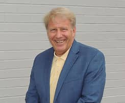 A headshot of an older man with blonde hair wearing a blue suit jacket and yellow shirt.