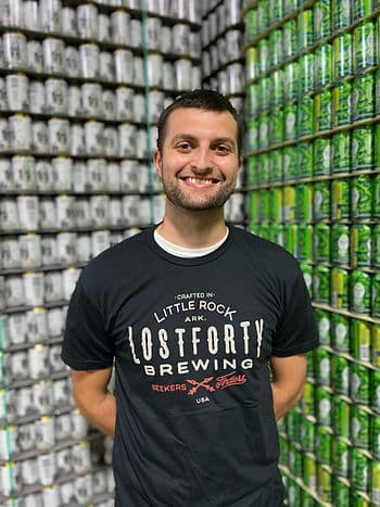 A headshot of a Lost Forty Brewing Company employee standing in front of a corner of two walls of beer, one white and one green.