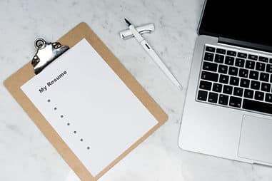 A clipboard with a blank sheet on it titled "My Resume" with blank bullet points. The corner of an Apple Macbook can be seen to the right of the clipboard ass well as a white fountain pen.