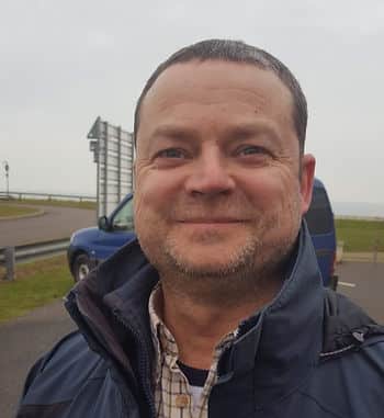 A headshot of a white man with thinning brown hair wearing a thick rain jacket over a plaid button-up.