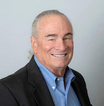 A headshot of an old man with gray hair wearing a black suit jacket and a blue shirt.