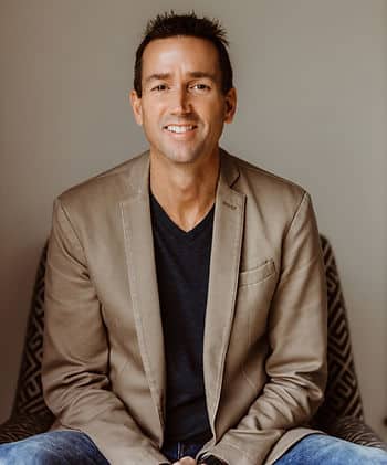 A headshot of a white man with black spiky hair wearing a tan suit jacket over a black T-shirt with jeans.