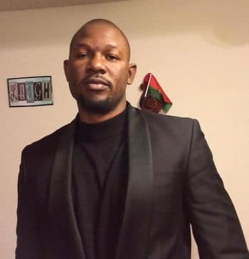 A headshot of a black man wearing a black suit and shirt.