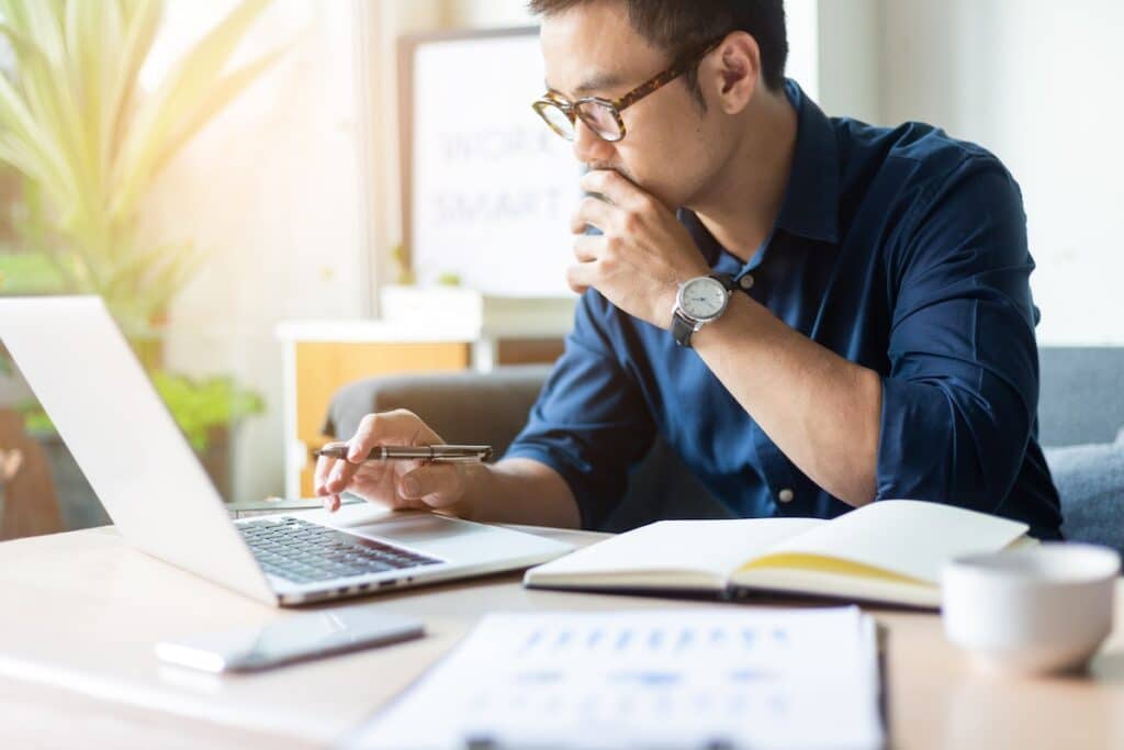 A man wearing glasses and a blue button up shirt sits at a table with his laptop and a notebook. He has one hand over his mouth and the other is holding a pen and typing as he works on internships.