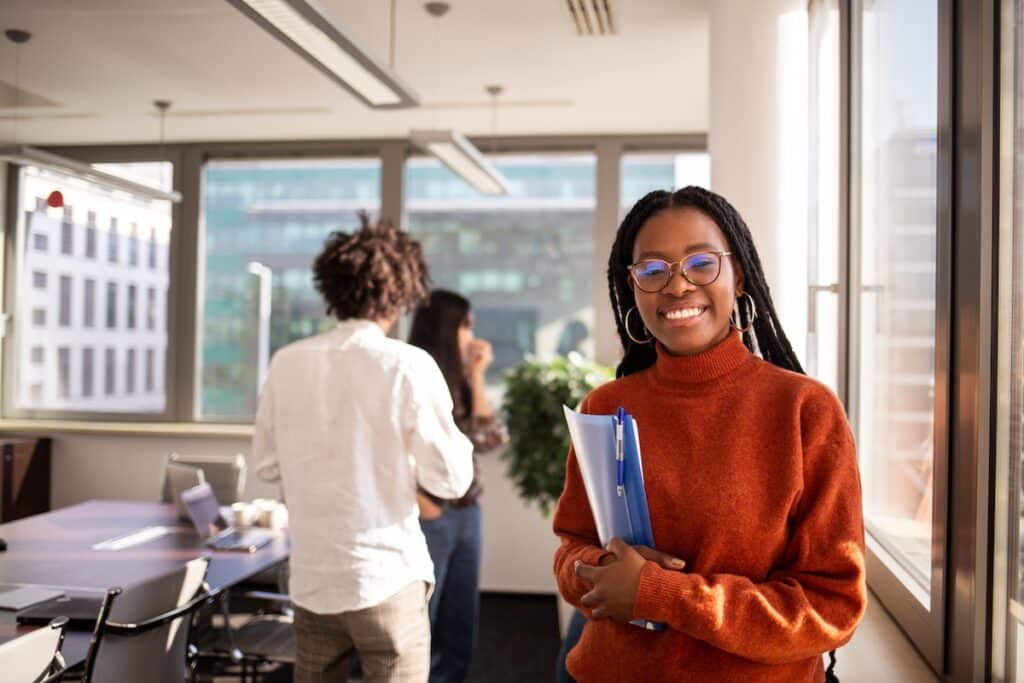 A girl in a registered apprenticeship program in the foreground is looking at the camera. She is wearing a burnt orange sweater and is holding a blue folder.