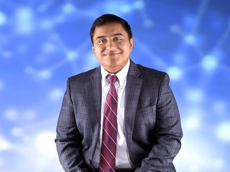 A headshot of an Indian man in a gray plaid suit with a red tie. The background is light blue with spots of light.